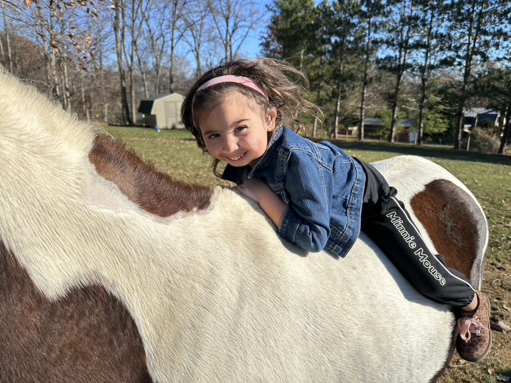 A smiling child lies across the back of a paint pony in the sunshine.