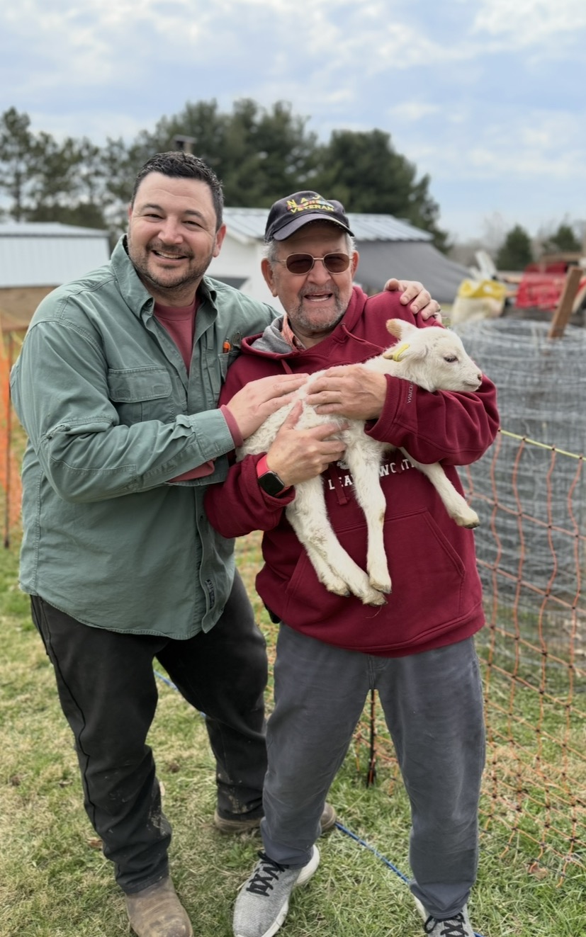 A veteran and a volunteer smile while cradling a newborn lamb at the ranch.